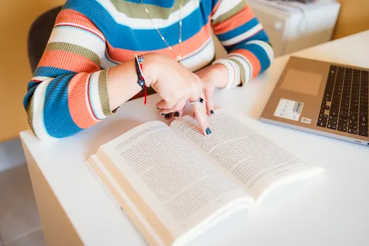 Mãos de Melissa apoiadas sobre um livro de estudo ao lado do notebook